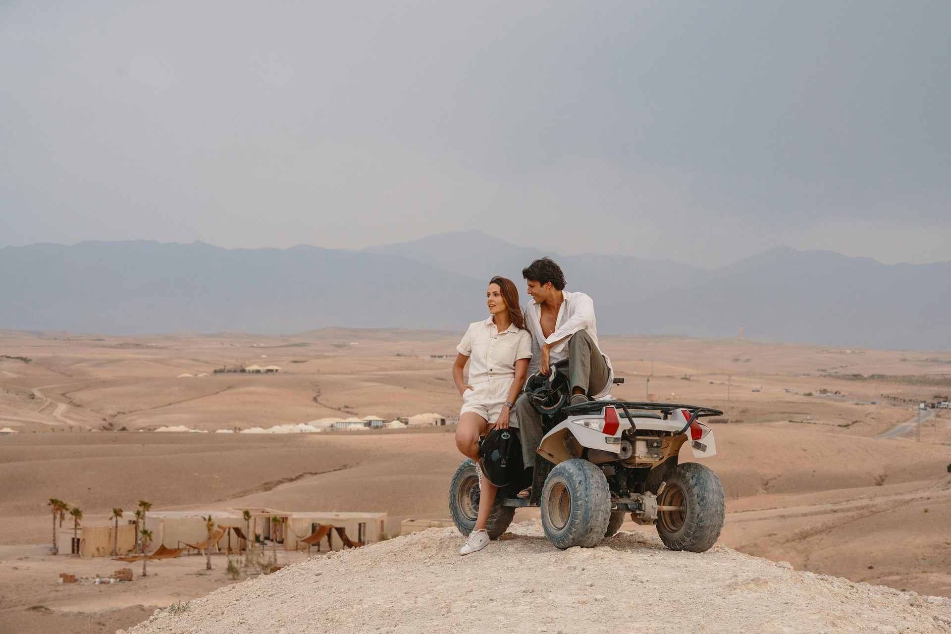 The bare rolling hills of the Agafay desert near Marrakech, with a tented camp and palm trees below and the Atlas Mountains on the horizon