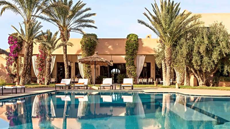 Pool reflecting date palms and bougainvillea in front of the villa facade and its sun loungers