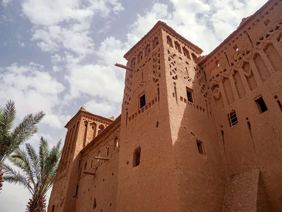 Uma alta torre de kasbah em terra compactada, as paredes superiores recortadas com padrões geométricos, vista de baixo contra o céu