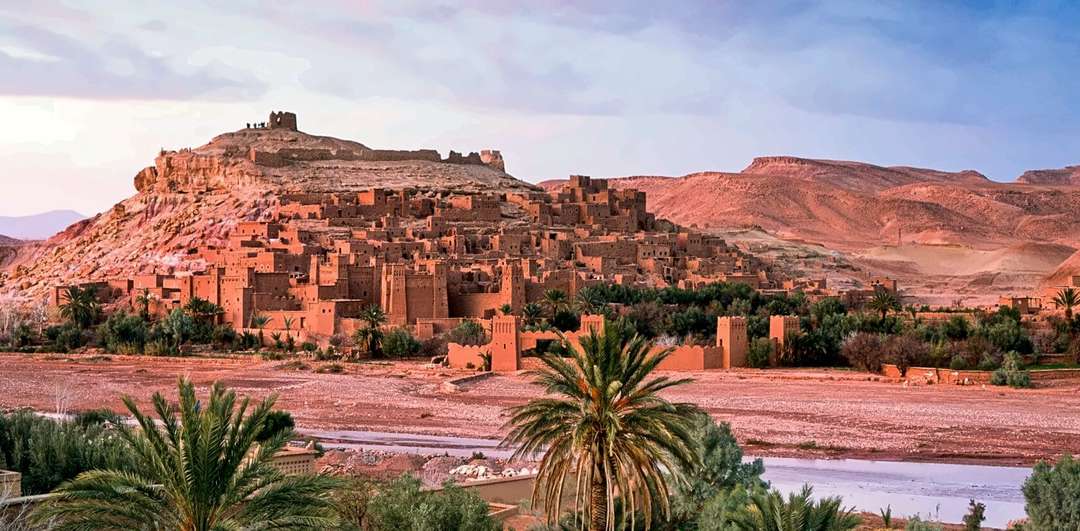 O ksar de Aït Ben Haddou ao crepúsculo, luz quente nas paredes de terra, com palmeiras e o leito seco do rio em frente