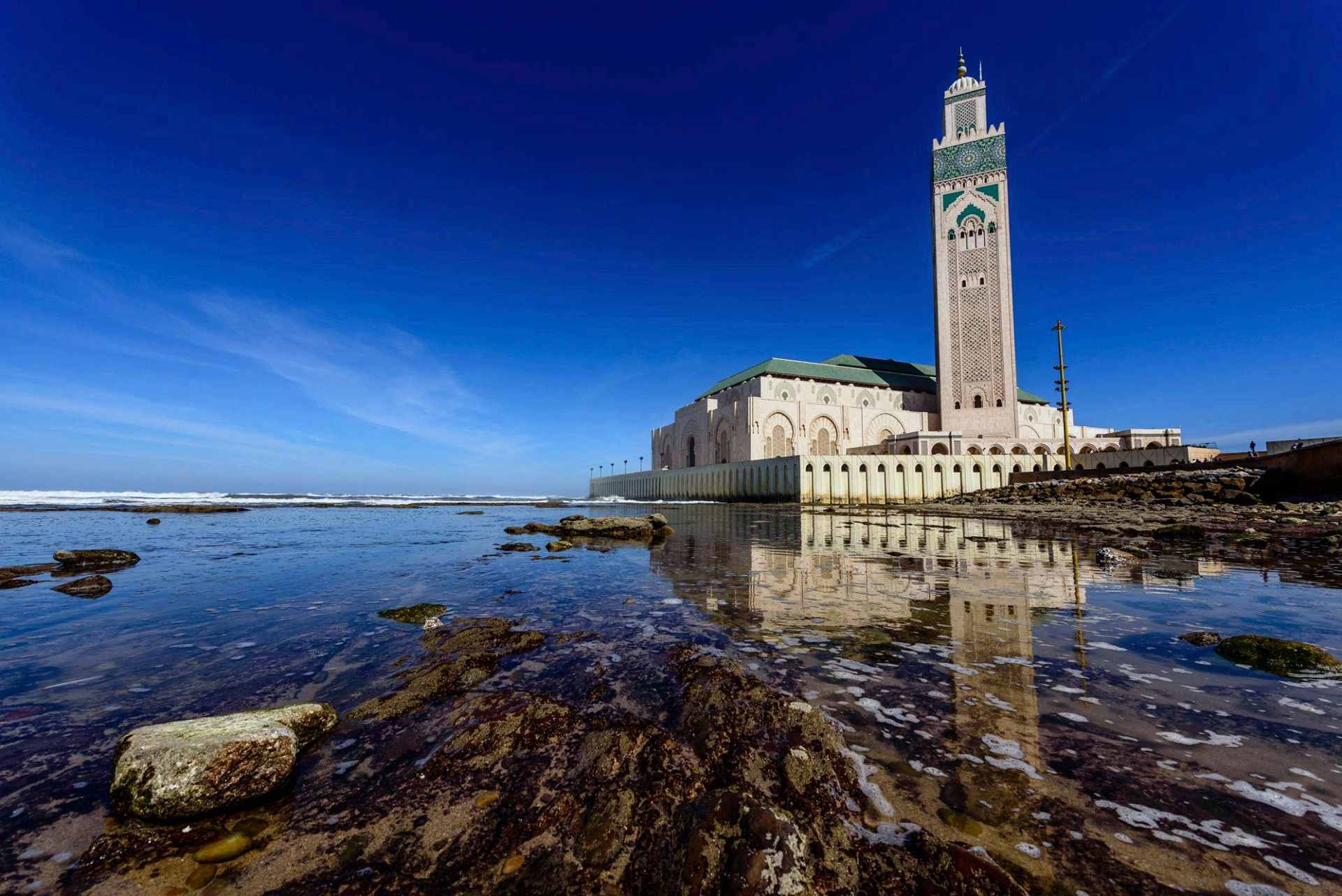 Die Hassan-II.-Moschee in Casablanca, von den Felsen bei Ebbe aus gesehen, ihr hohes Minarett und die Ufermauer spiegeln sich unter tiefblauem Himmel im Wasser