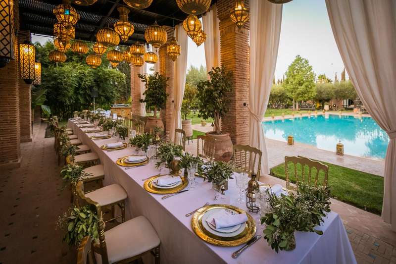 Un lungo tavolo apparecchiato per una festa sotto lanterne marocchine, su una terrazza piastrellata accanto a una piscina illuminata al crepuscolo