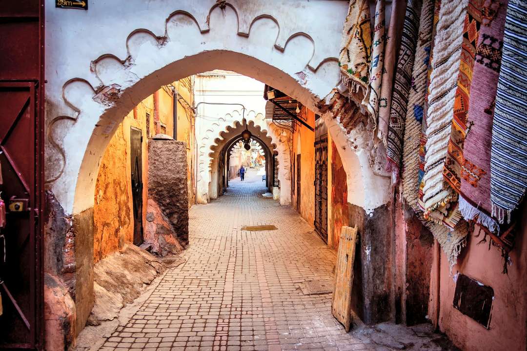 Un vicolo dei souk della medina di Marrakech che corre sotto archi smerlati, tappeti e coperte appesi lungo il muro