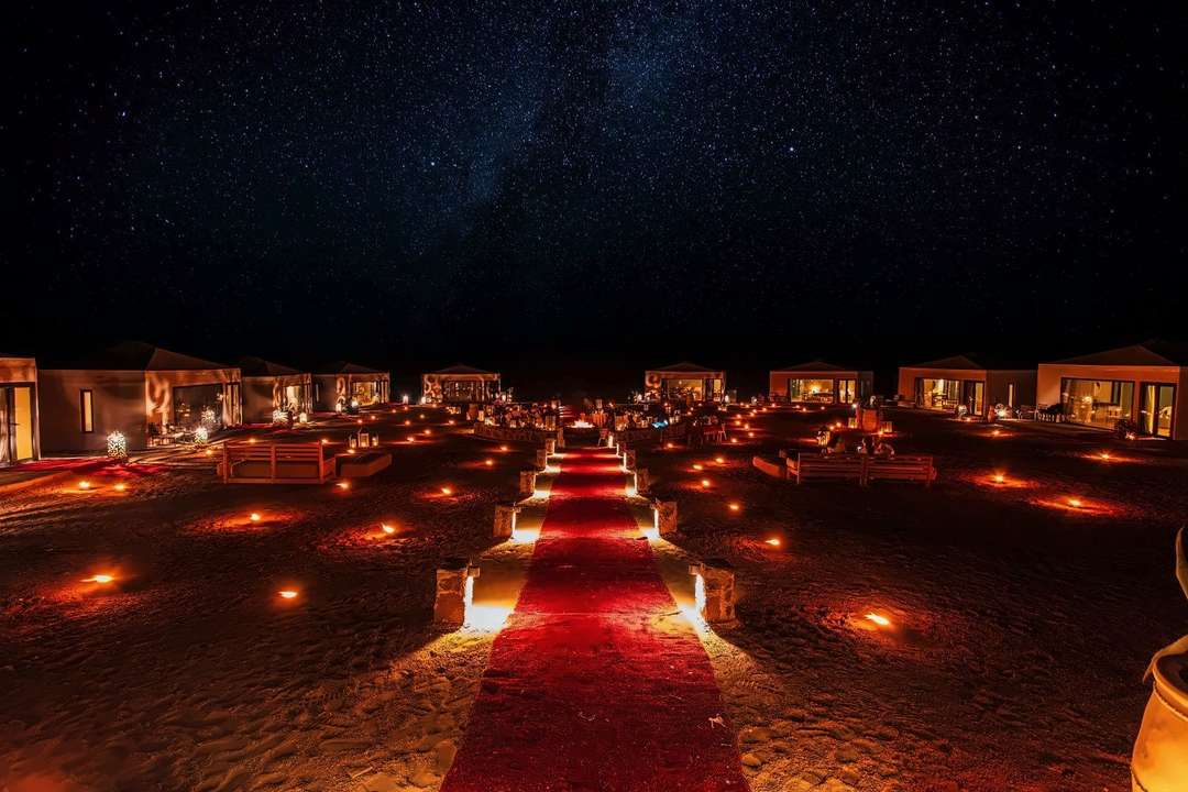 Um acampamento no deserto de noite, tendas iluminadas e lanternas ao longo de um caminho de tapetes sob um céu cheio de estrelas