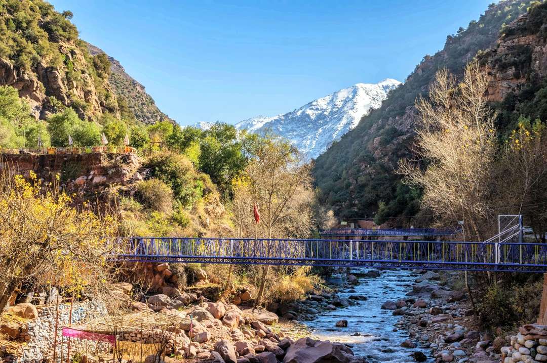 Uma passarela metálica azul sobre o rio Ourika, esplanadas de restaurante com guarda-sóis na margem e um cume nevado por trás