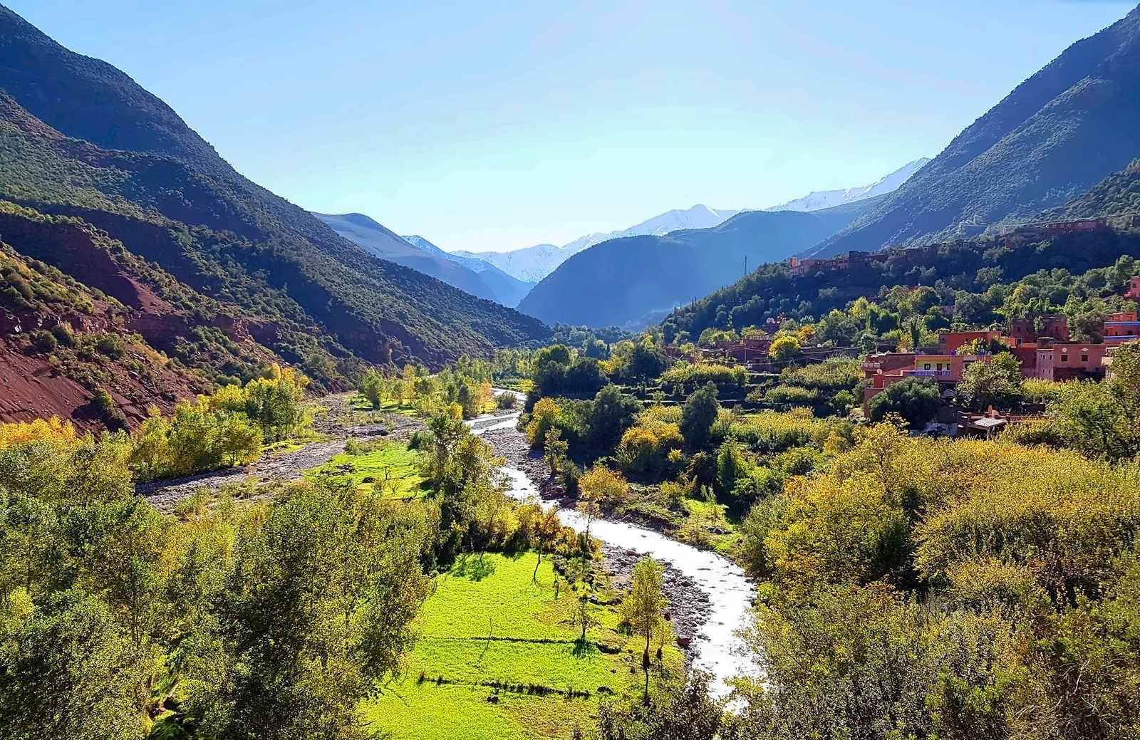 O rio Ourika a correr entre margens verdejantes, uma aldeia na encosta e o Alto Atlas nevado ao longe