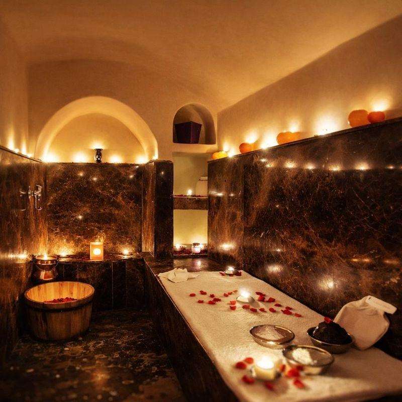 Die Behandlungsplatte eines Hammams in Marrakesch im Kerzenlicht, mit Rosenblättern, Schalen mit schwarzer Seife und Rhassoul-Lehm und einem Holzeimer neben dem Marmor