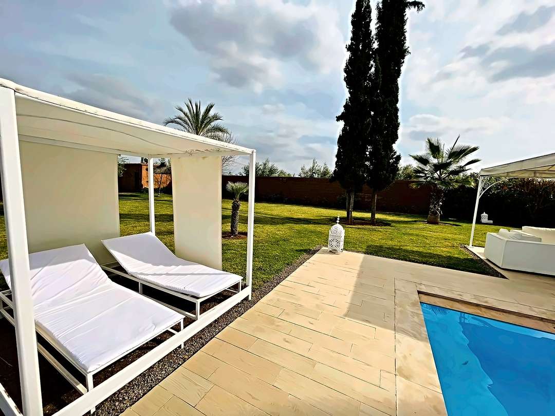 Letto a baldacchino e due chaise longue imbottite a bordo piscina, tra cipressi e palme