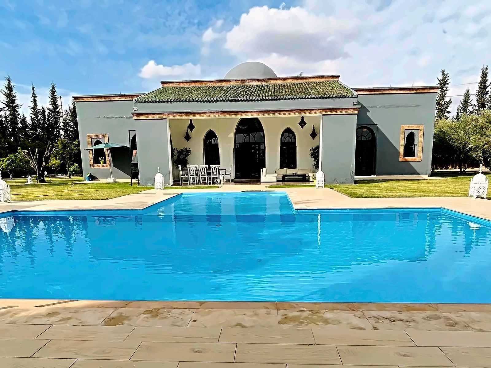 Grande piscina davanti alla villa a un solo piano, con tetto di tegole verdi, cupola e terrazza ad archi
