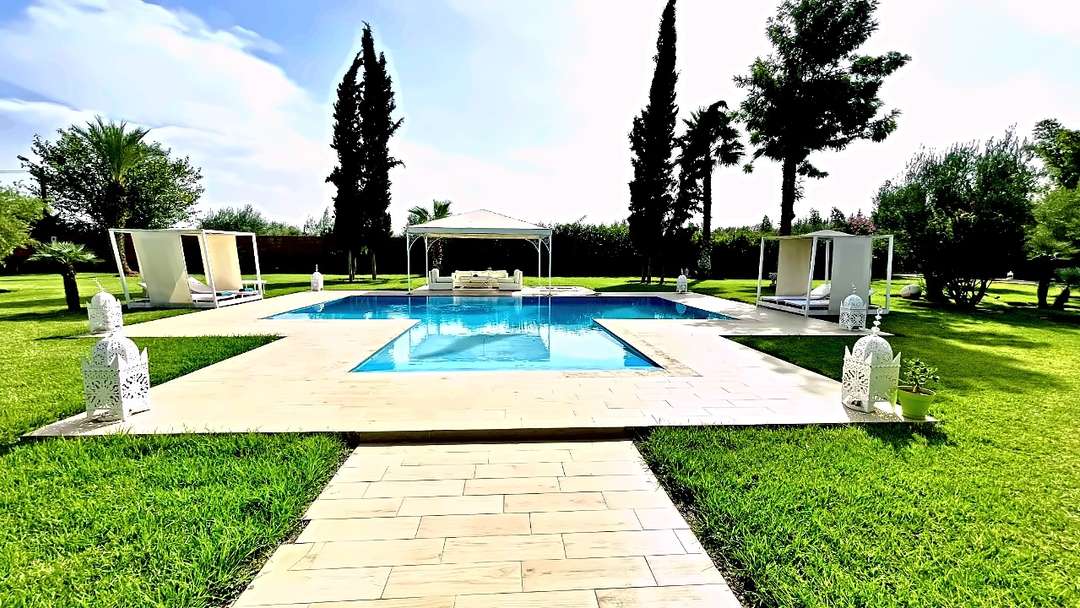 Piscina a croce su un ampio prato, con gazebo bianco, due letti a baldacchino e lanterne marocchine