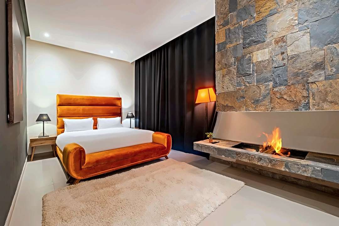 Schlafzimmer mit Kopfteil und Bank in orangem Samt gegenüber einem Steinkamin mit brennendem Feuer