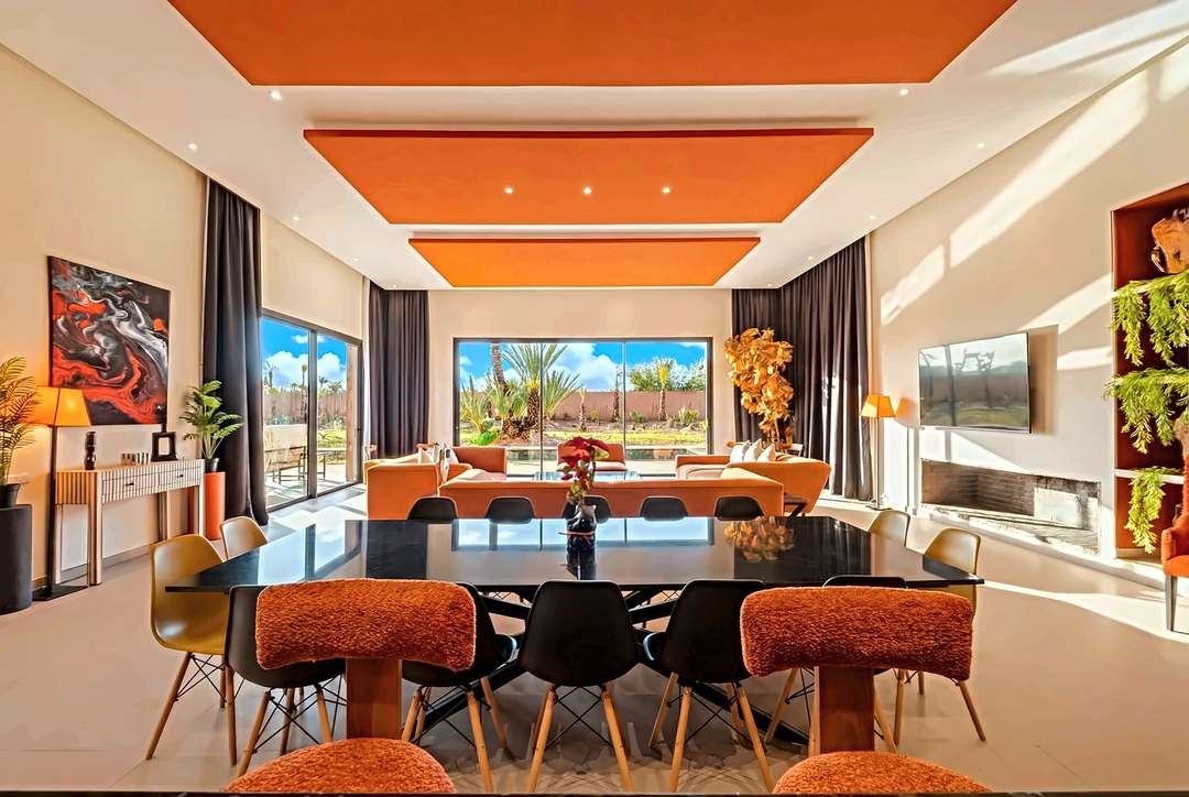 Esstisch für zwölf Personen und orange Sofas unter orangen Deckenpaneelen, mit Glastüren zum Garten