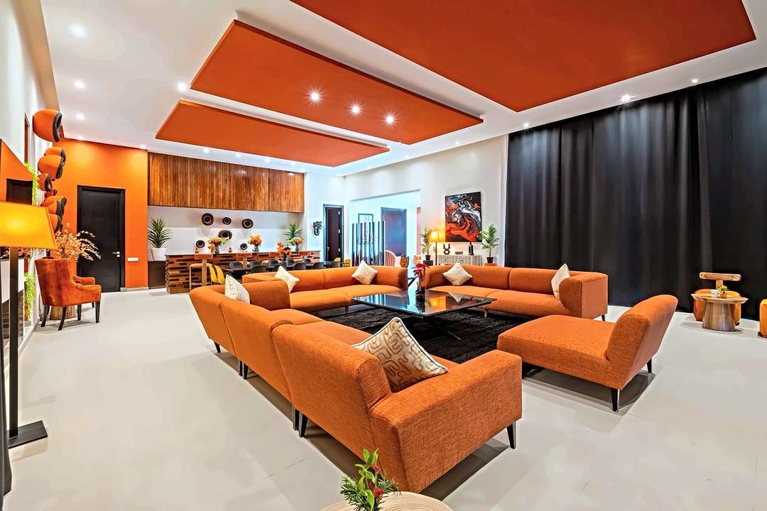Großes Wohnzimmer mit orangen Sofas um einen schwarzen Tisch, orangem Deckenpaneel und Bar dahinter
