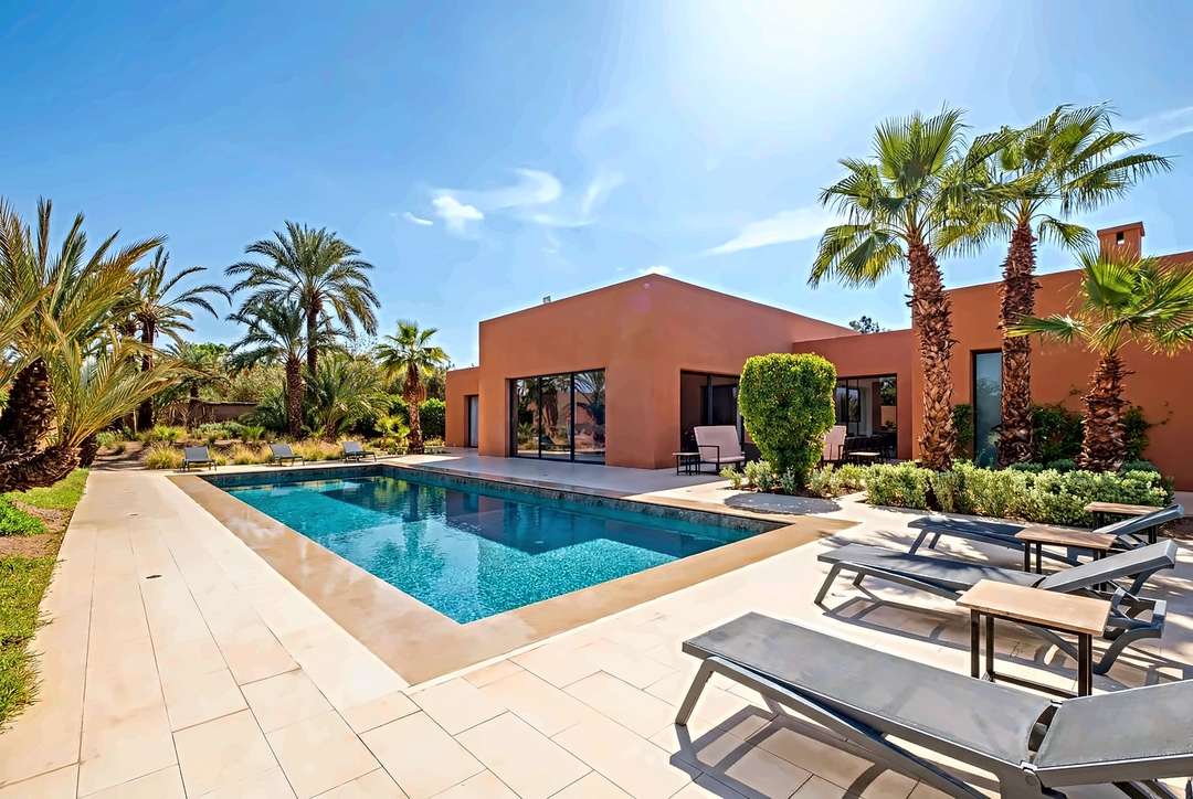 Poolterrasse mit grauen Sonnenliegen und Beistelltischen, Fächerpalmen und der Villa in Terrakotta in voller Sonne