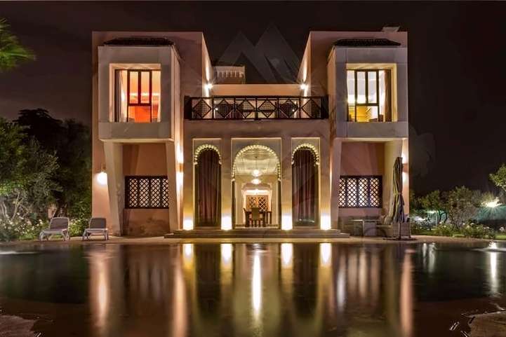Villa su due piani illuminata di notte, con porte ad arco e balcone riflessi nella piscina