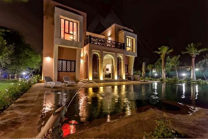Piscina con getto di fontana di notte, davanti alla villa illuminata e alle sue chaise longue