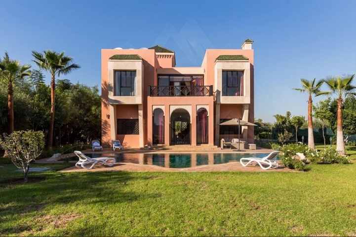 Villa rosa su due piani, con porte ad arco e balcone, rivolta verso una piscina e un ampio prato con palme